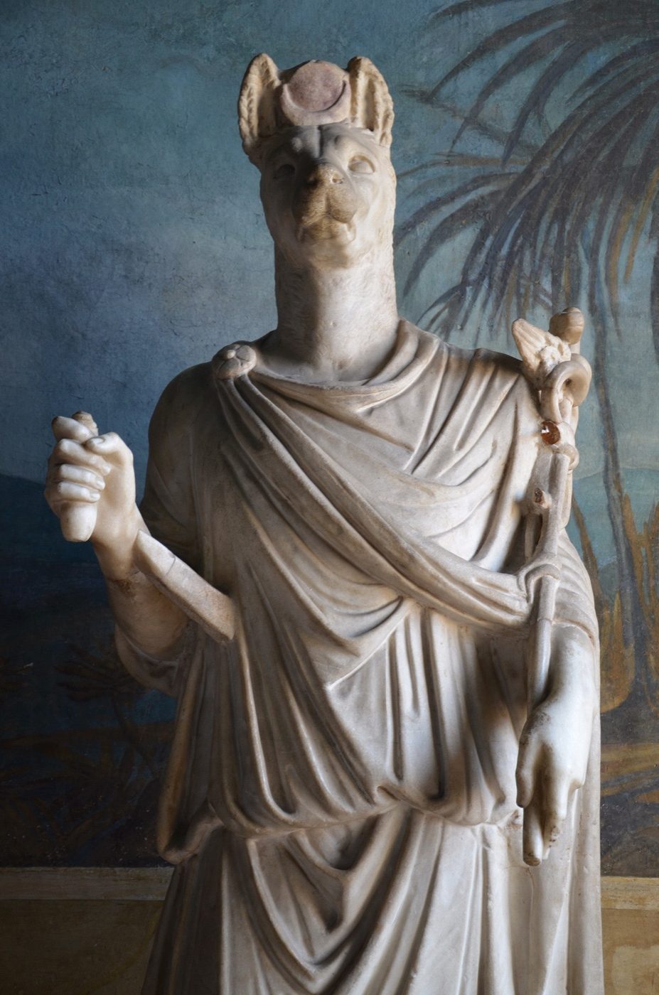 Roman-period marble statue of Anubis from Anzio, Italy, 1st-2nd century AD, dressed in a toga in the Roman style but shown with a jackal head, holding a sistrum and a caduceus borrowed from Hermes, at the Vatican Museums.