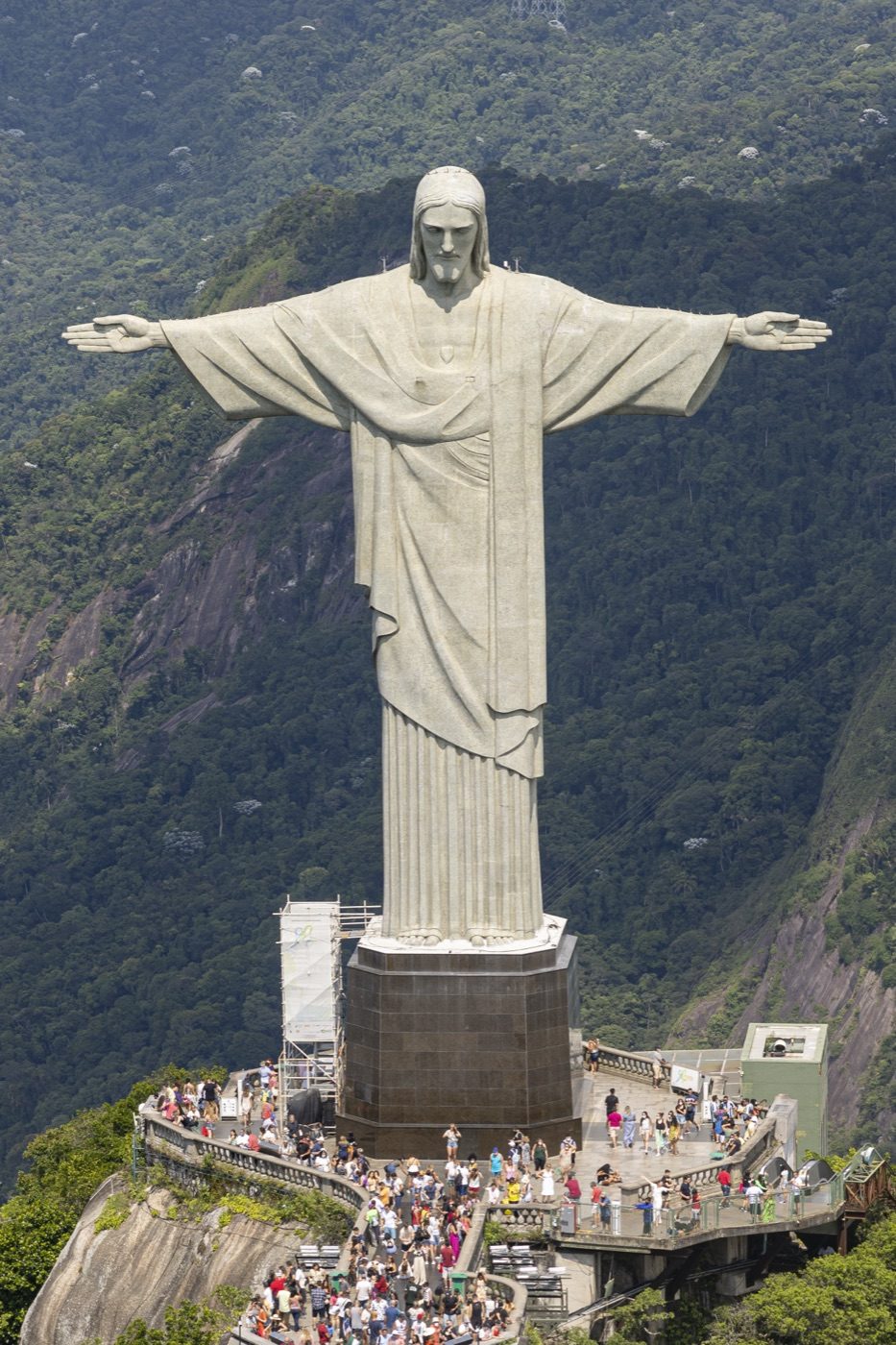 Aerial photograph of the Christ the Redeemer statue on Corcovado mountain overlooking Rio de Janeiro, with the viewing platform, its crowds of visitors, and forested mountainside visible below.