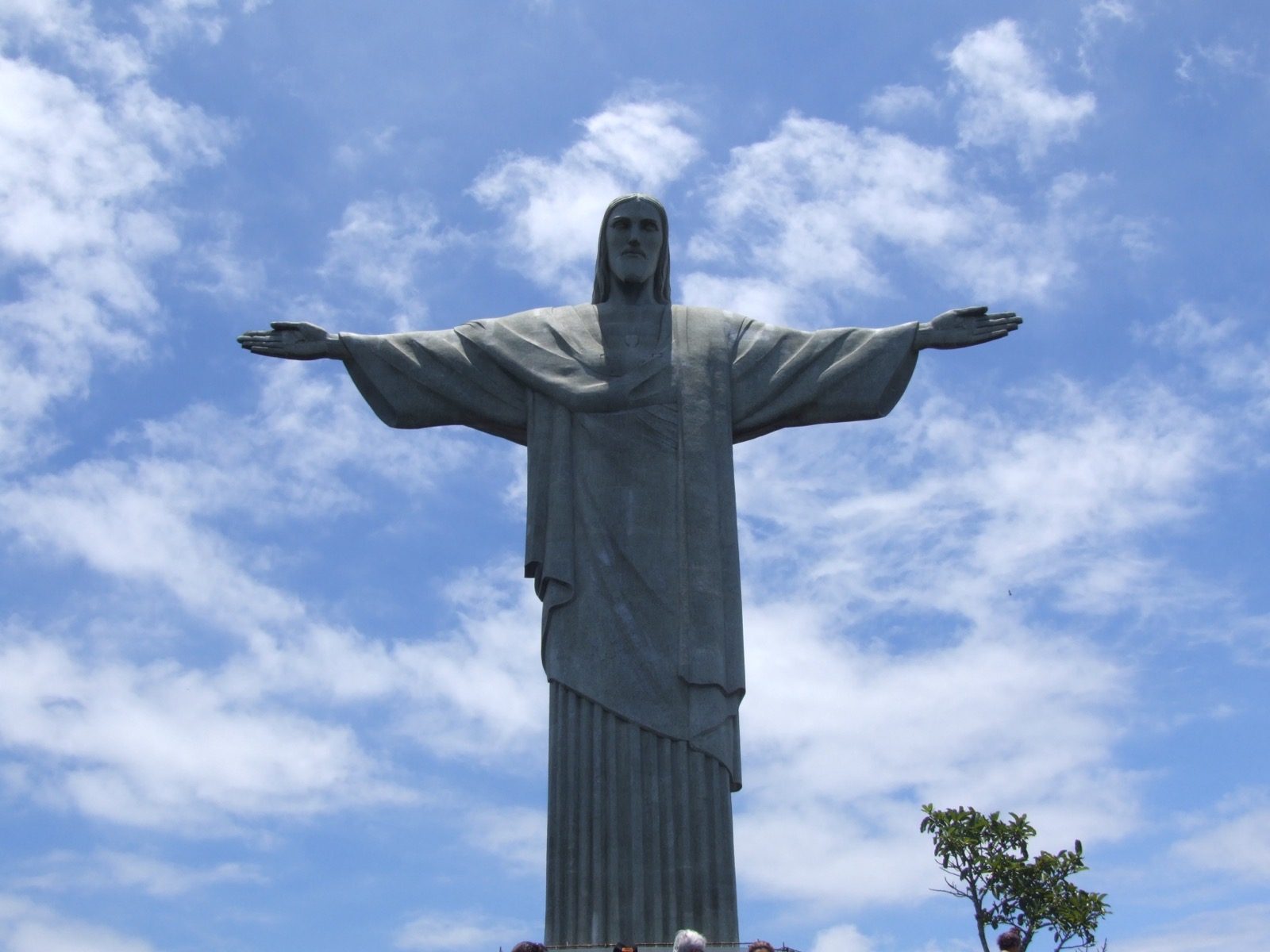 The Christ the Redeemer statue standing atop Corcovado mountain with its arms outstretched against a partly cloudy sky, overlooking the forested mountainside below.