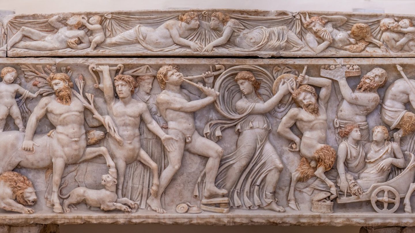 A detail from a Roman marble sarcophagus showing a Dionysian procession, with a bearded, goat-legged satyr and other mythological figures carved in high relief.