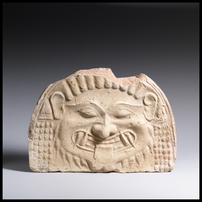 A terracotta gorgoneion antefix from an ancient Greek temple roof, showing the wide-eyed, wide-mouthed face of a Gorgon used as a protective architectural ornament, dated about 580-570 BCE.